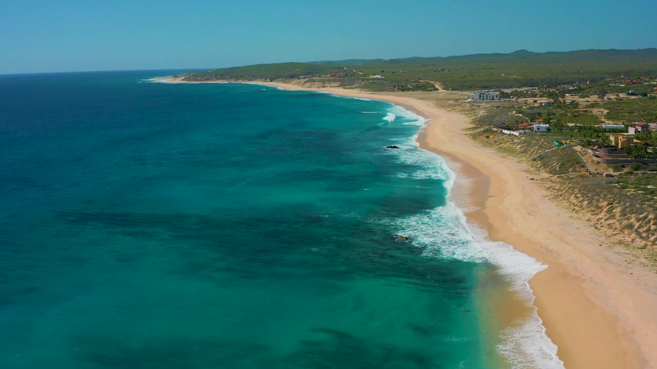A high coastal view in El Cardoncito shows white wave bands meeting the sand, low dunes and scrub behind the beach, scattered buildings inland, and no visible people.