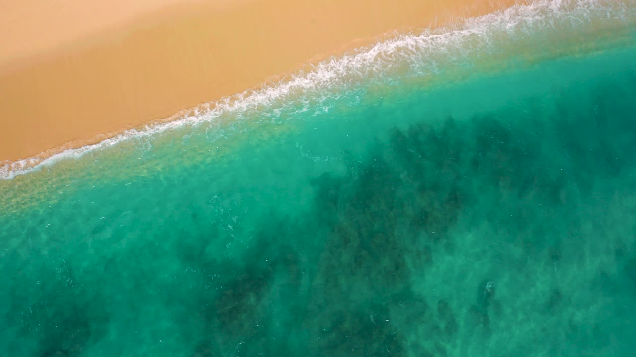 Overhead view of El Cardoncito shoreline with tan sand at the top, a thin white surf line, clear turquoise water below, and no visible people.