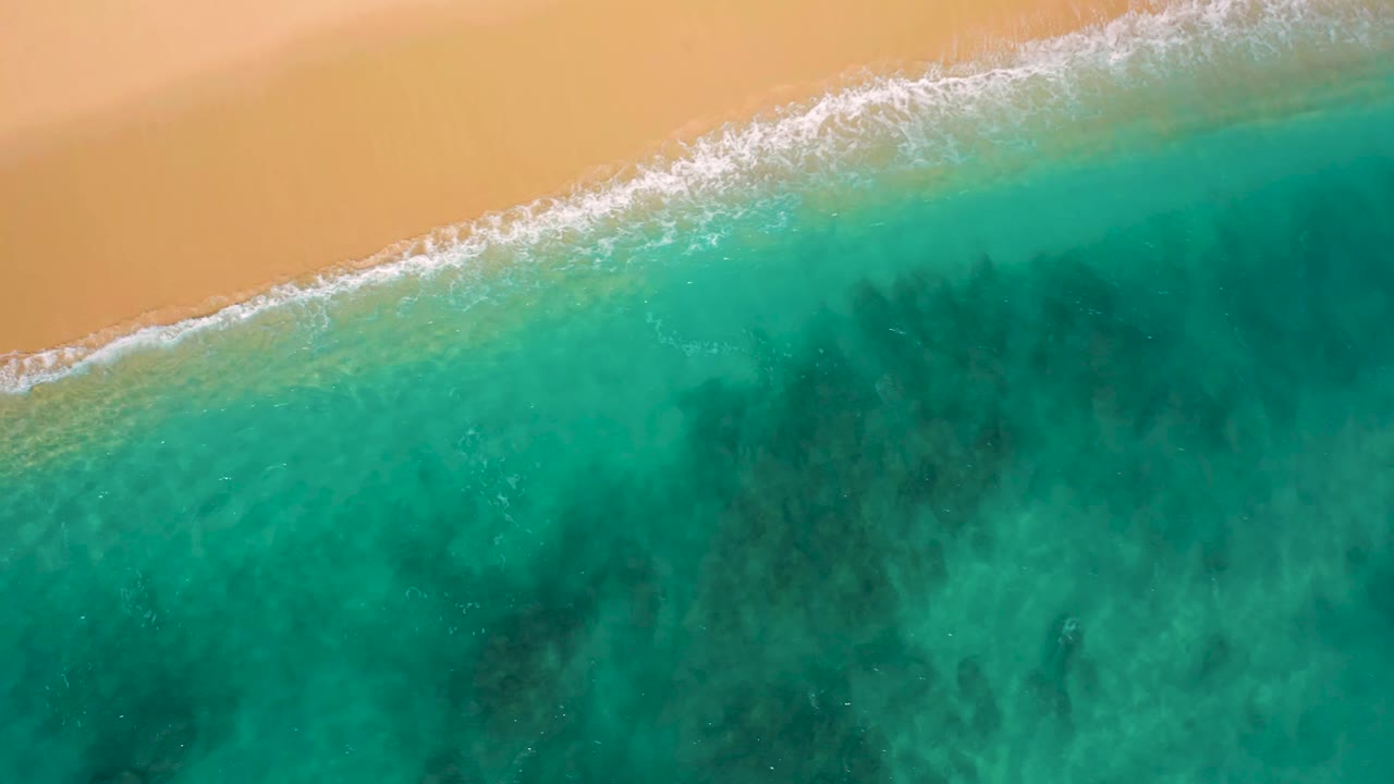 Aerial frame over El Cardoncito with the surf line slightly farther across the sand edge, green-blue shallows filling the lower frame, and no visible people.