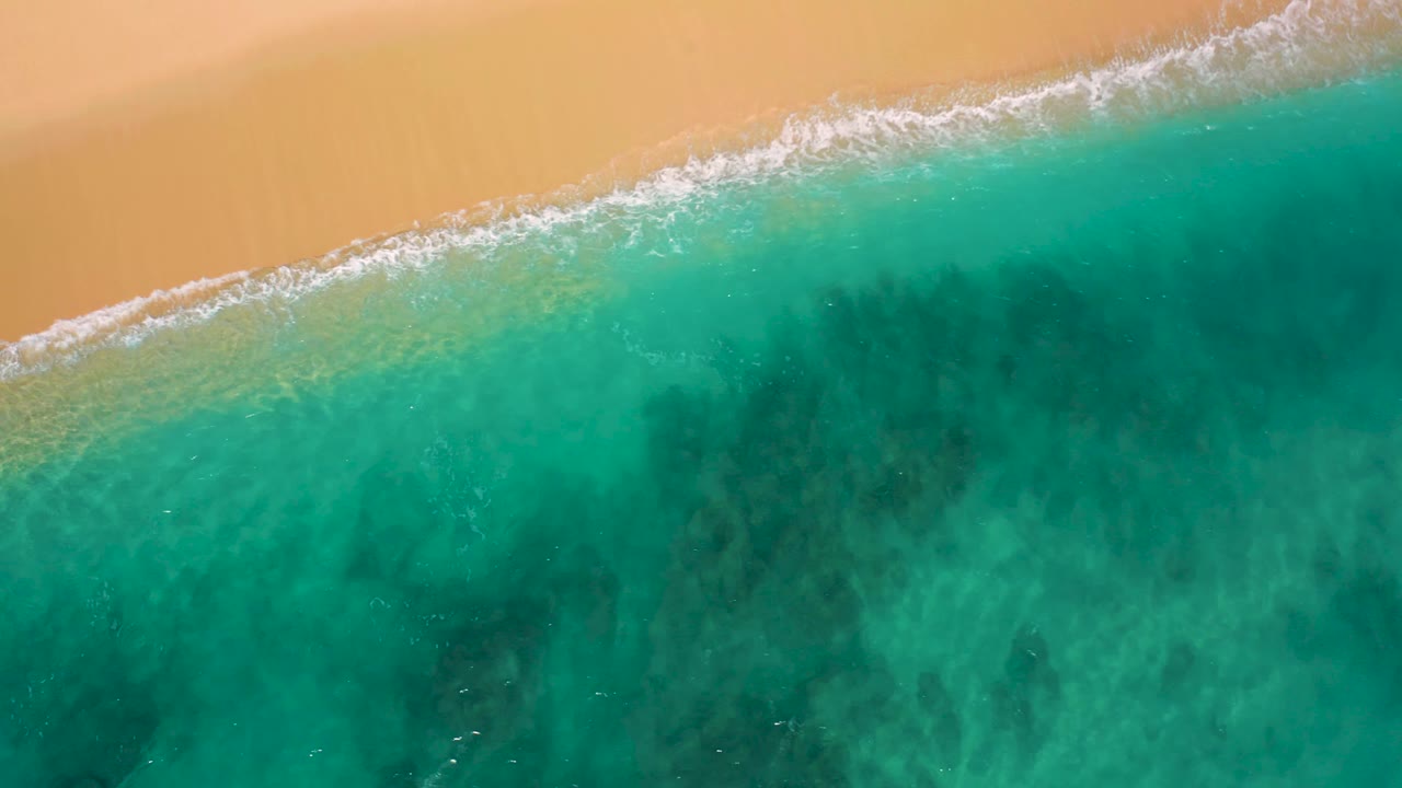 Top-down El Cardoncito shoreline view with foamy wash crossing diagonally, pale sand above it, darker green water below, and no visible people.