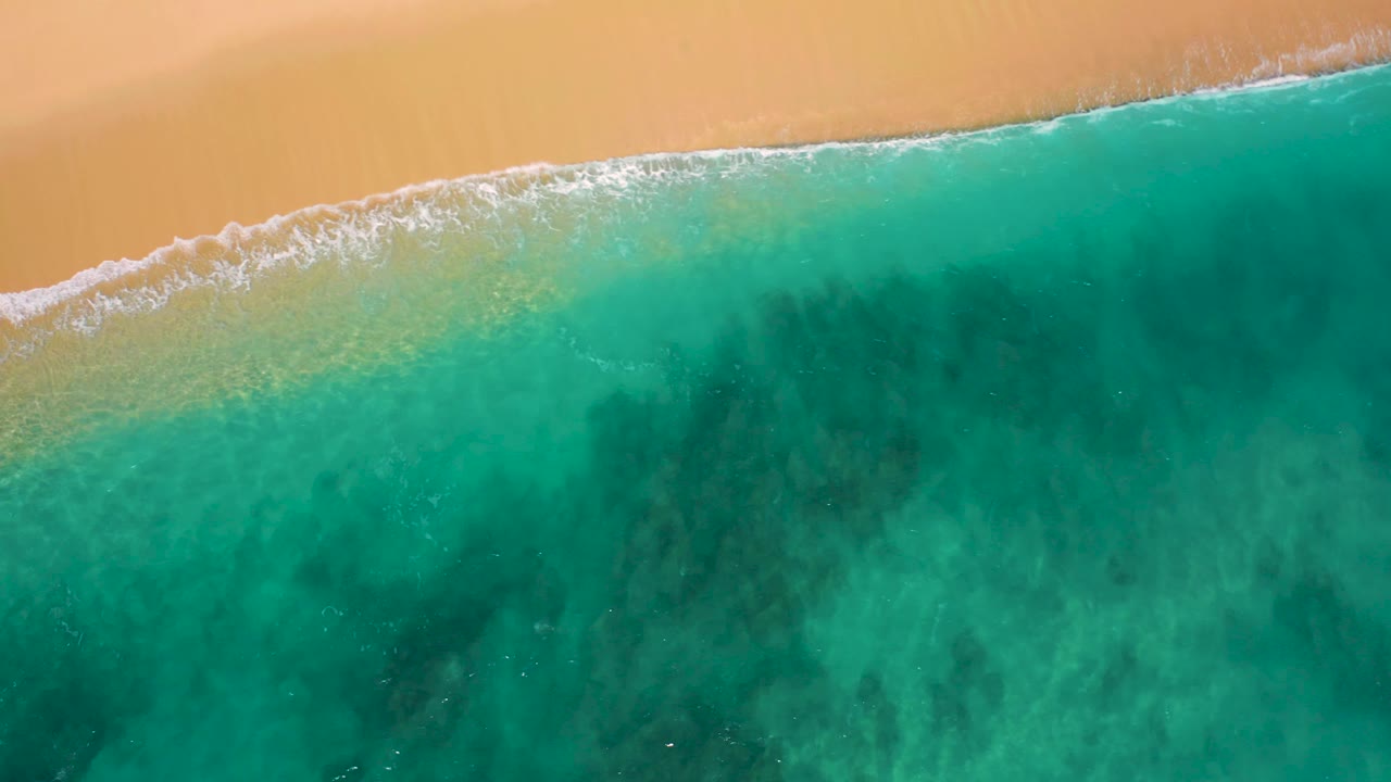 Overhead frame from El Cardoncito showing a broader wash of white foam over tan sand, turquoise water spreading through the center, and no visible people.