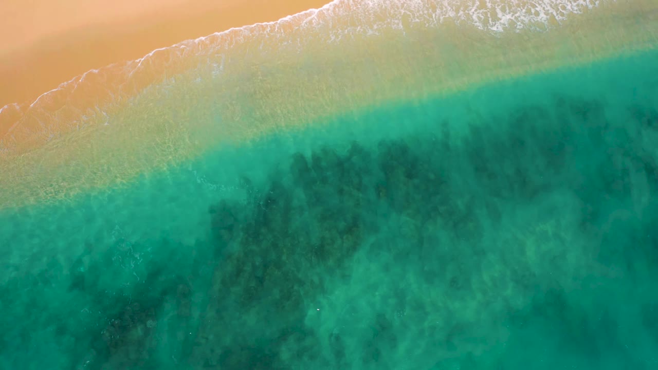 Aerial view in El Cardoncito with foamy wave edges sliding along the upper shoreline, green-blue shallows below, darker submerged areas, and no visible people.