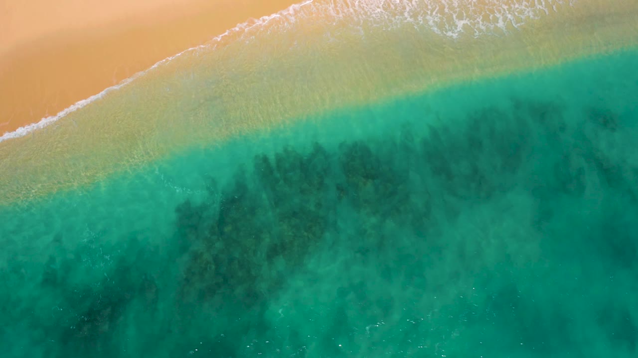 Aerial view over El Cardoncito showing a wider sweep of turquoise water below a pale sandy beach, with faint white foam along the shore and no visible people.