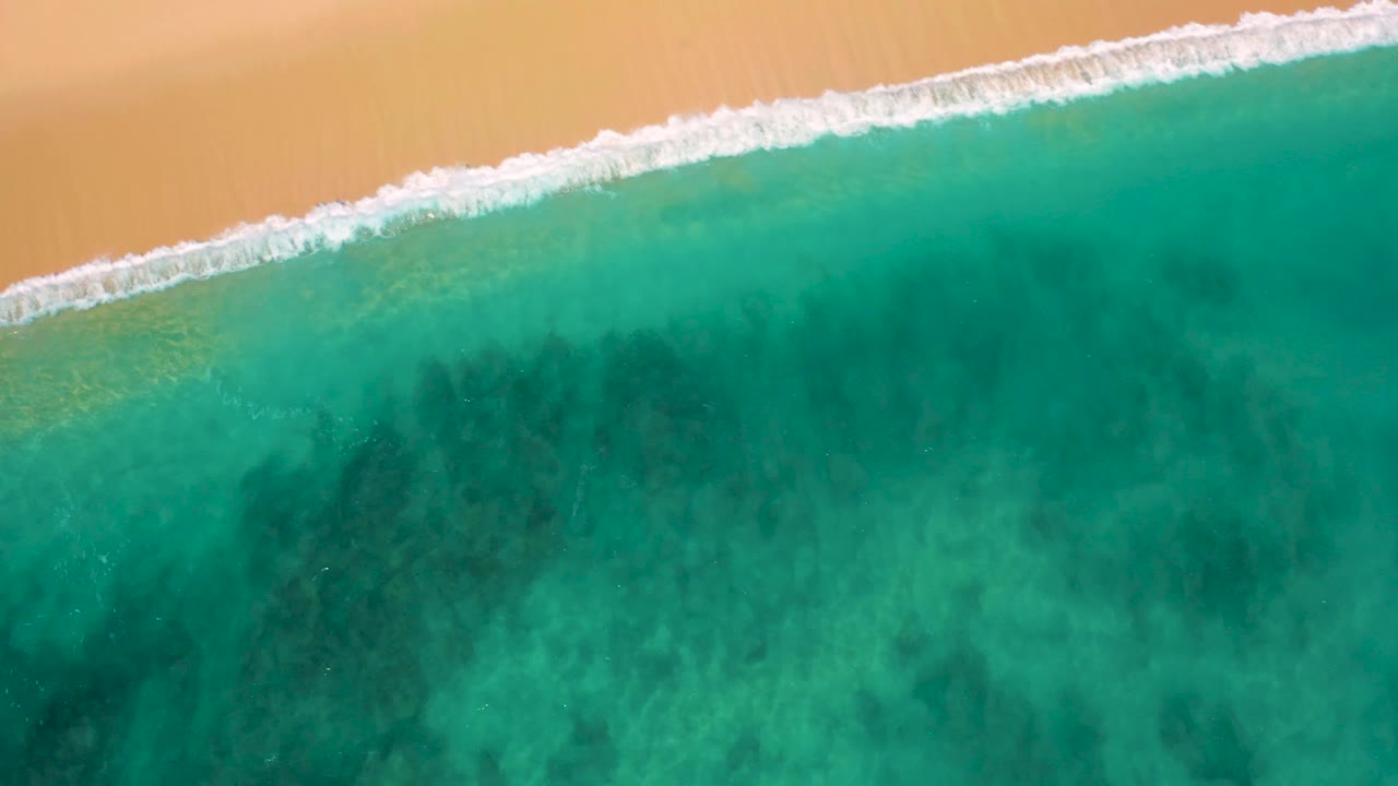A broader band of white foam reaches the El Cardoncito sand while clear green-blue water fills most of the frame, with no visible people.