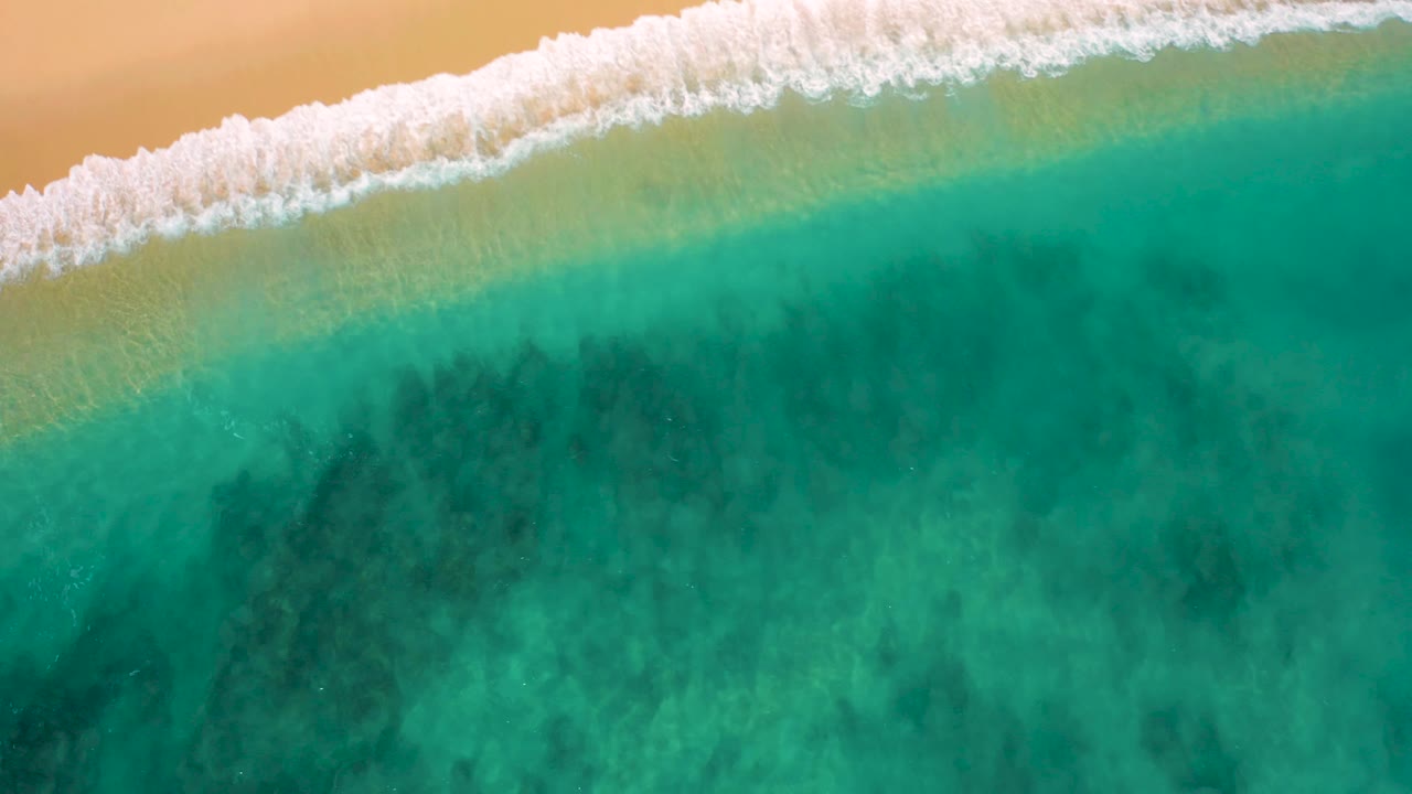Overhead view of El Cardoncito surf with white foam across the upper left, a pale shallow-water band, teal water below, and no visible people.