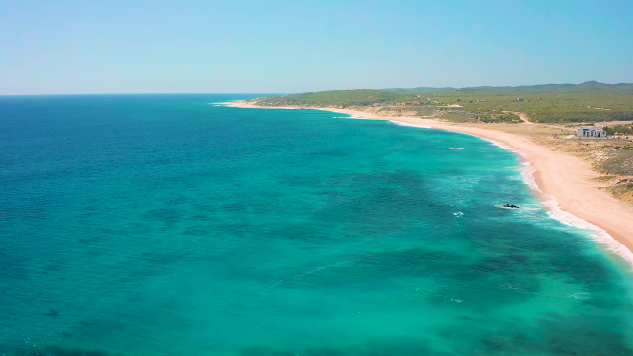 Wide aerial frame of El Cardoncito showing blue-green sea across most of the image, a narrow surf edge on the sandy shore, inland vegetation, distant hills, and no visible people.