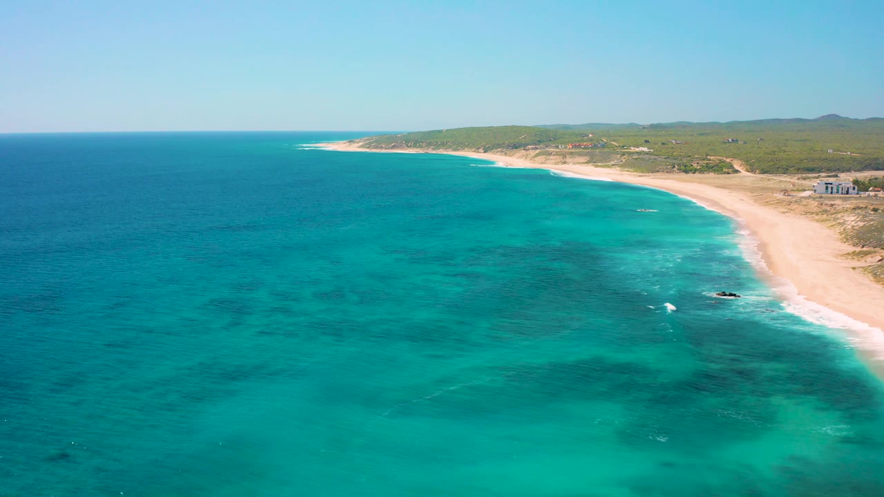 Aerial view over El Cardoncito where darker patches in the water lead toward a small rocky area near the surf, with the sandy beach and low green inland terrain visible, and no visible people.