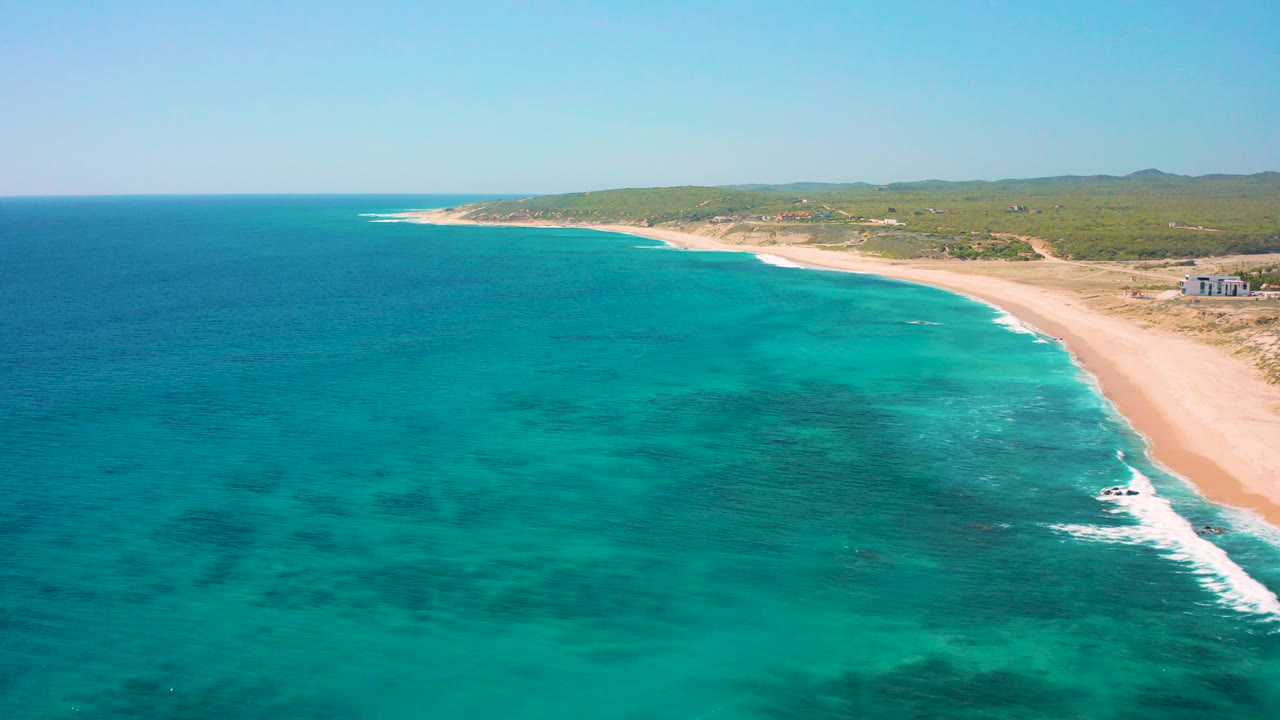 Opening aerial frame over El Cardoncito, Baja California Sur, with turquoise water, a curving sandy beach, low green inland terrain, and no visible people.