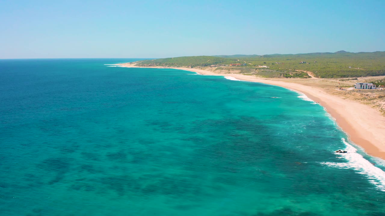 At 0:02, the El Cardoncito shoreline remains in a wide aerial composition, with darker underwater patches visible through turquoise water and no visible people on the beach.