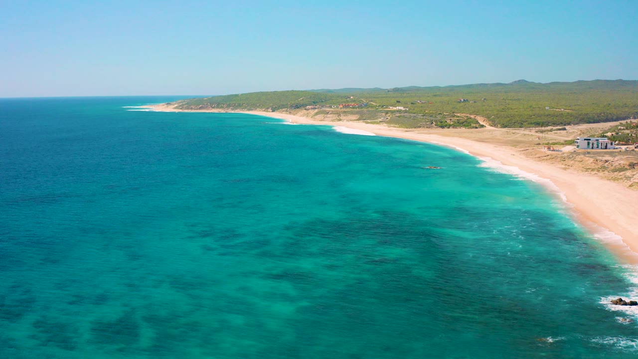Aerial frame over El Cardoncito, Baja California Sur, with blue-green water in the foreground, a pale sandy beach on the right, low vegetation inland, and no visible people.