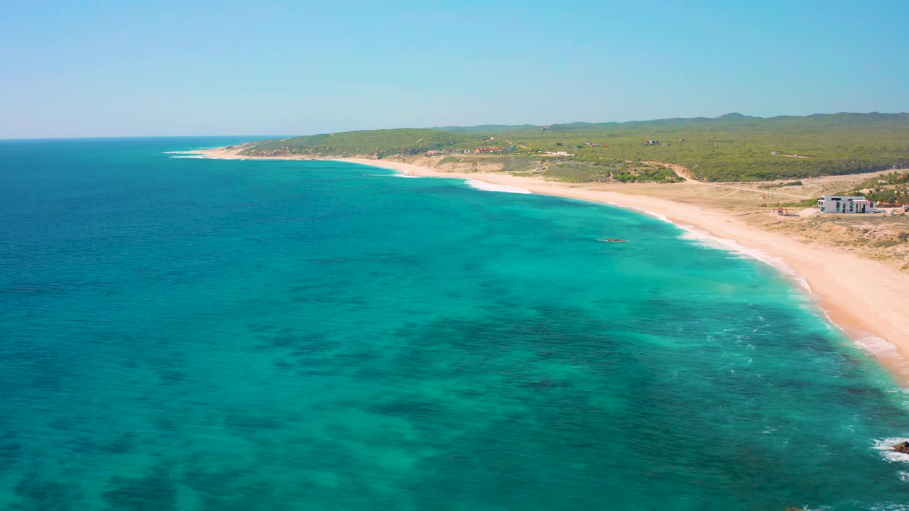 High aerial frame over El Cardoncito with darker patches under the blue-green water, a narrow surf line along the beach, low green terrain inland, and no visible people.