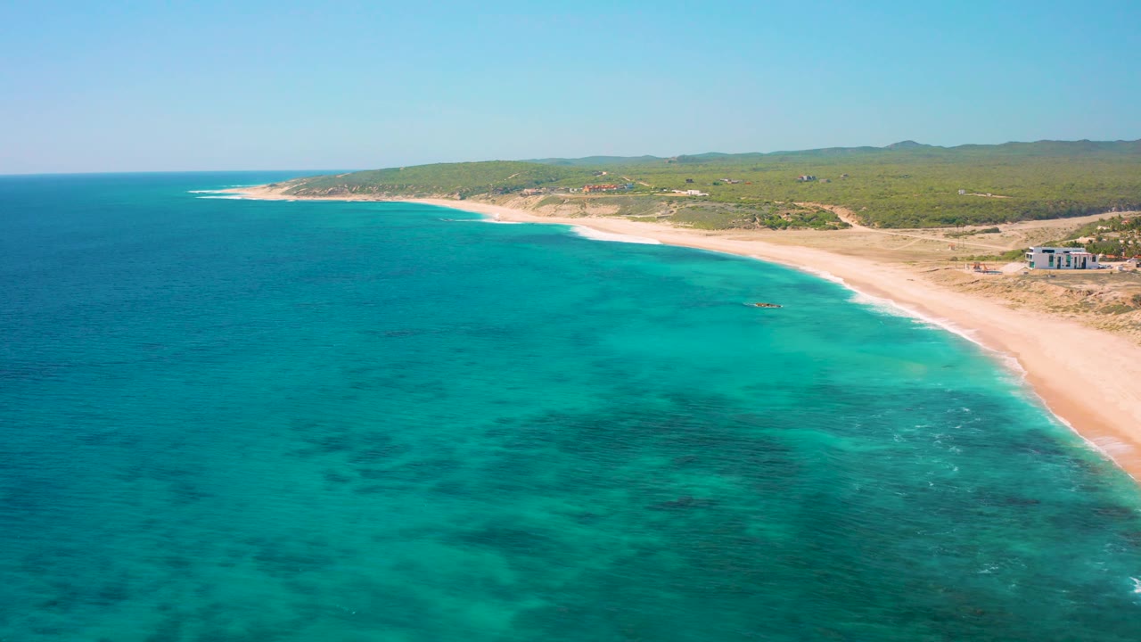 Aerial coastline view in El Cardoncito where open blue water occupies the left side, lighter shallows meet the sandy beach on the right, and no visible people appear on shore.