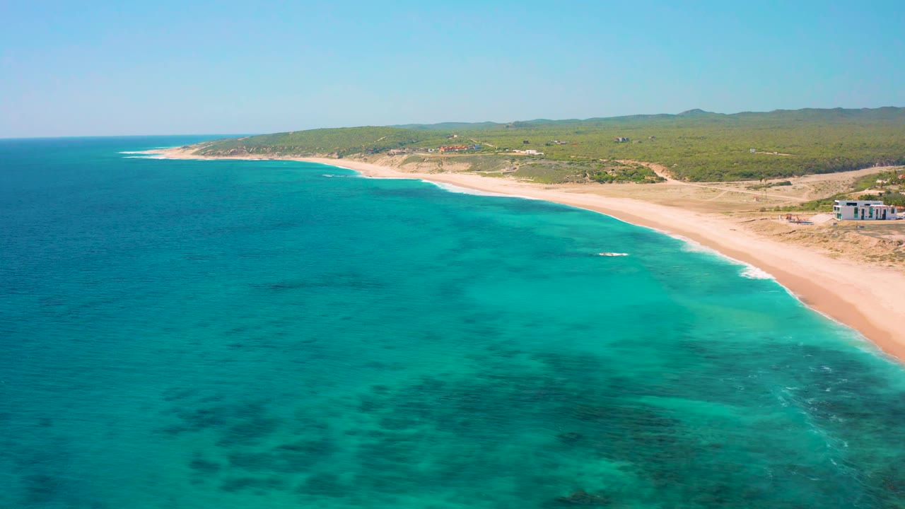 Aerial frame at El Cardoncito showing clear turquoise sea, a long pale beach, scattered dark underwater patches, inland scrub, distant hills, and no visible people.