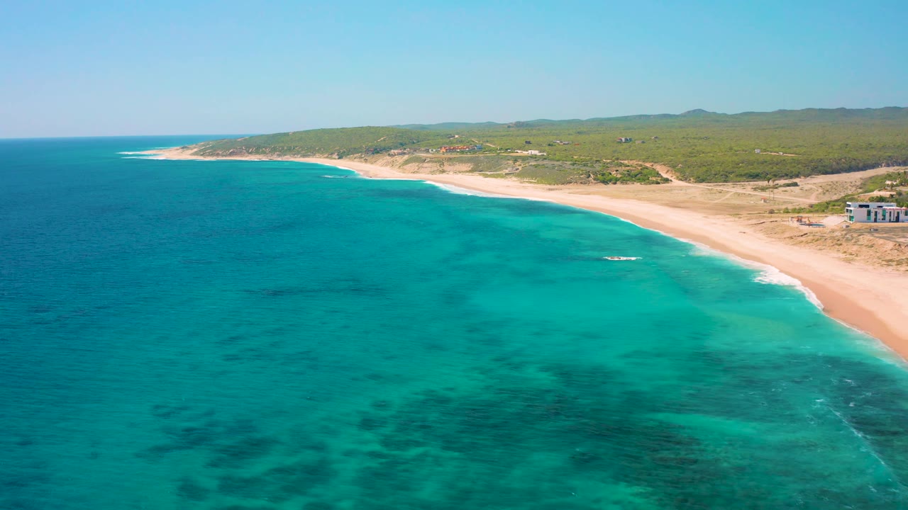 Wide aerial view of El Cardoncito with turquoise shallows spreading across the foreground, a pale beach on the right, scrubland behind it, a few small buildings, and no visible people.