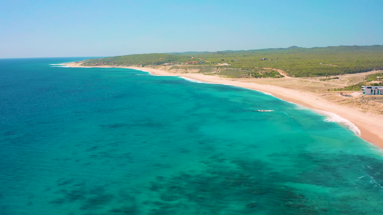 Offshore aerial view near El Cardoncito with the ocean filling most of the frame, a pale sand beach along the right, low vegetation beyond the shore, and no visible people.