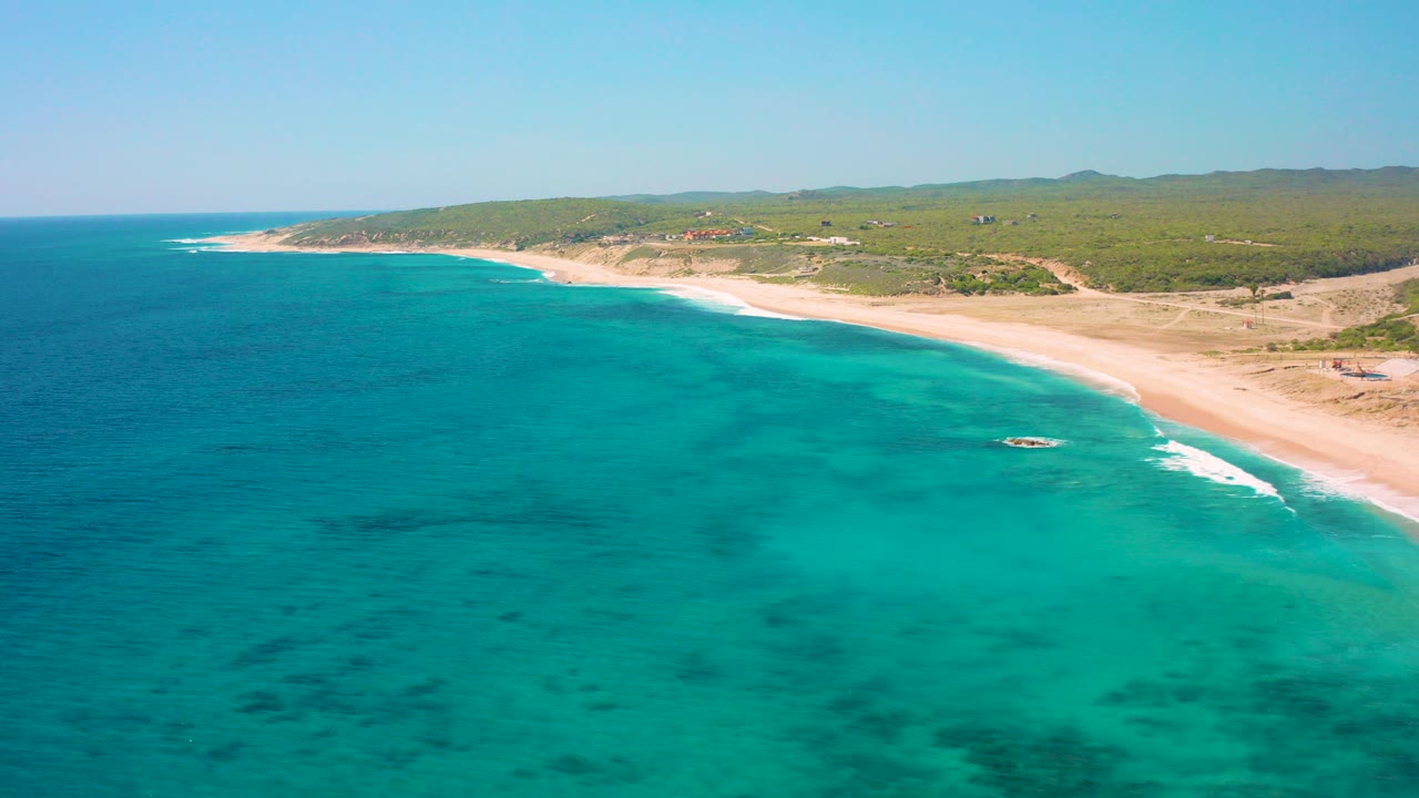 Aerial view over El Cardoncito, Baja California Sur, showing turquoise ocean in the foreground, a pale sandy beach curving across the frame, inland dunes and green vegetation, and no visible people.