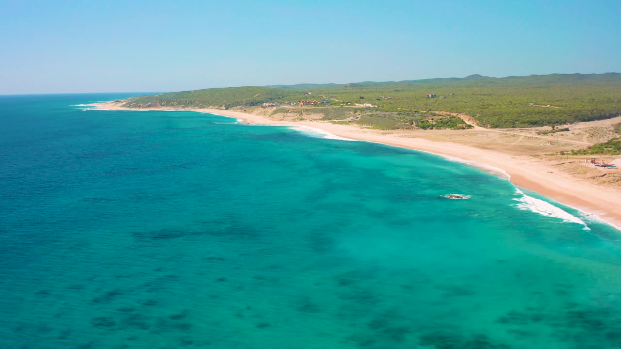 Aerial view at El Cardoncito with blue-green ocean spread across the foreground, a long sand beach along the right side, low brush inland, distant hills, and no visible people.