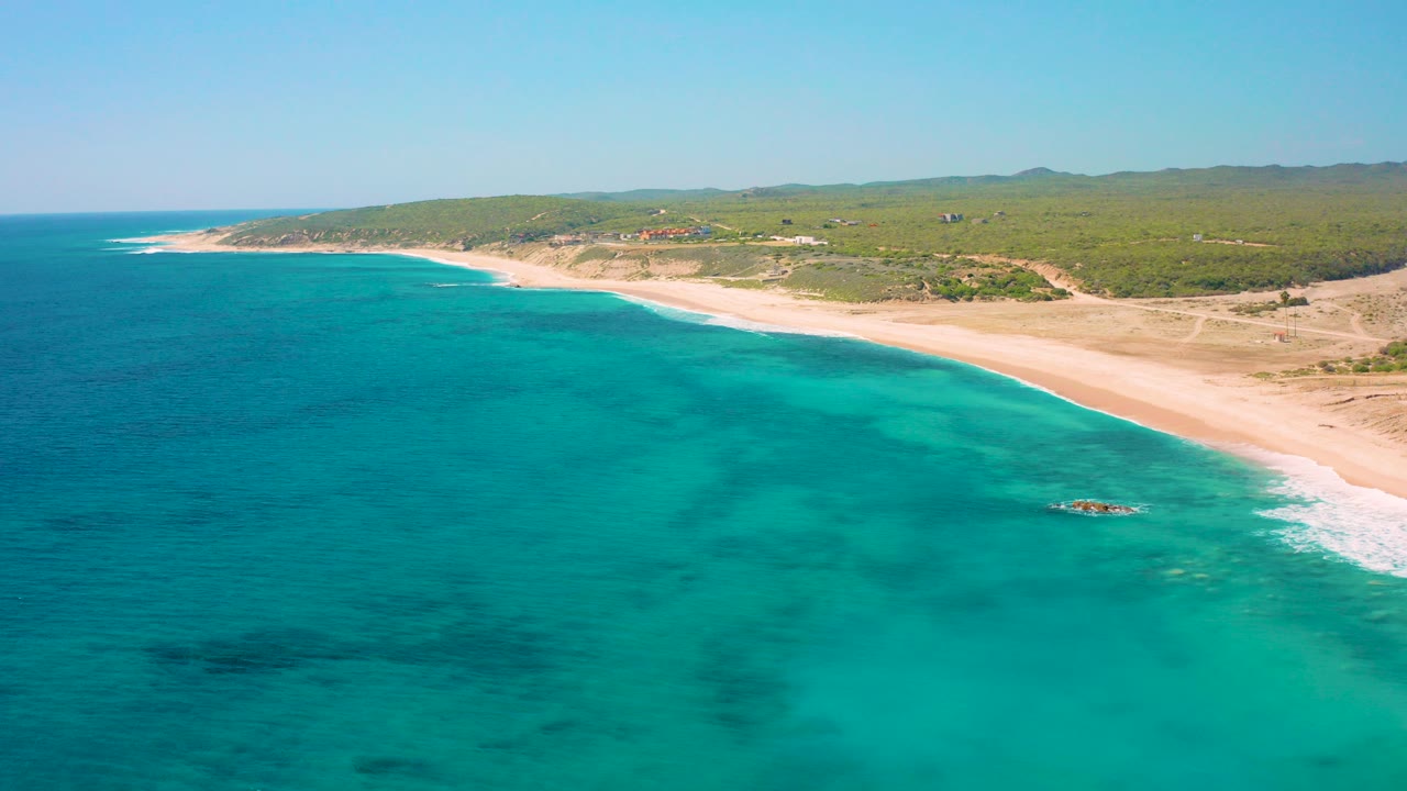 High aerial view over El Cardoncito, Baja California Sur, with turquoise water in the foreground, a long sandy beach at right, inland scrub beyond the shore, and no visible people.