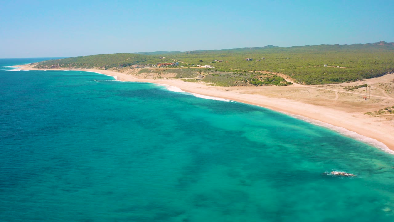 Opening aerial frame over El Cardoncito, Baja California Sur, with turquoise water in the foreground, pale sand along the shore, green scrubland beyond, distant hills, and no visible people.