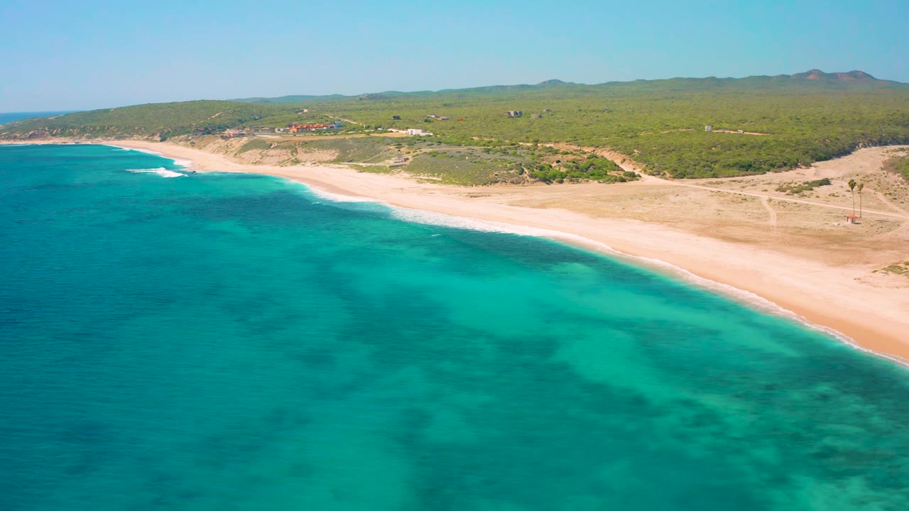 Aerial view at El Cardoncito showing clear blue-green shallows, a long sandy shoreline, inland scrub vegetation, distant hills, and no visible people.