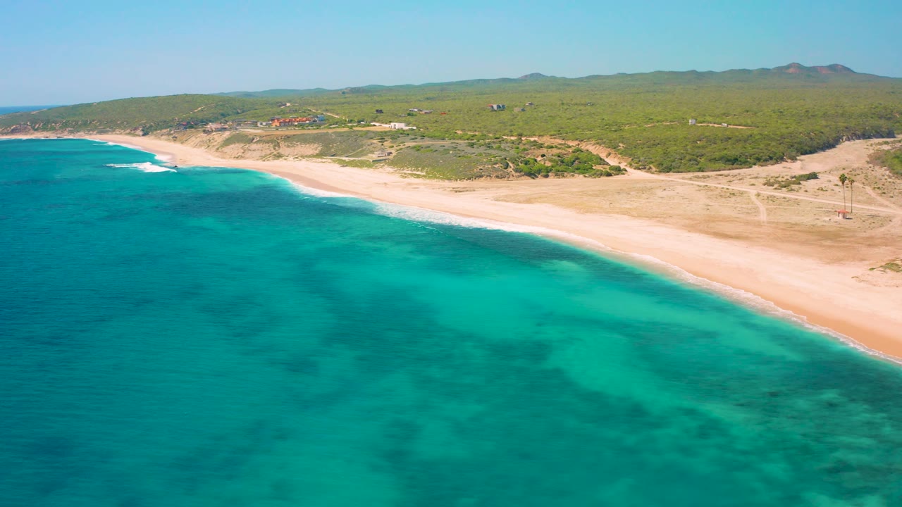 El Cardoncito aerial frame with darker reef-like patches in turquoise water, a clean sand strip, scrub-covered land behind it, low hills on the horizon, and no visible people.