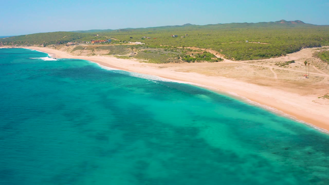 Wide aerial view of El Cardoncito with turquoise sea in front, pale sand and faint dirt tracks on the beach area, green inland terrain, distant hills, and no visible people.
