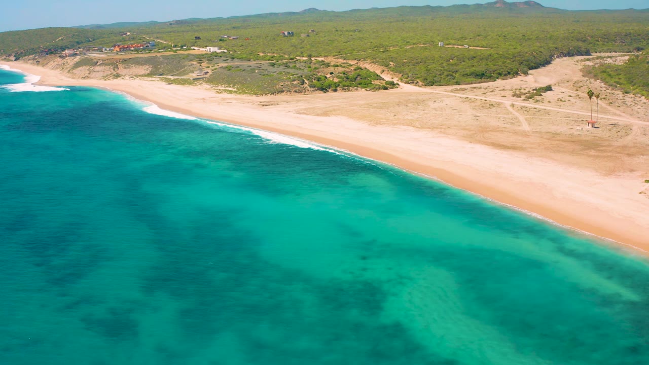 Aerial view in El Cardoncito with more open water filling the lower frame, pale sand along the upper diagonal, scrub-covered inland ground, small distant buildings, and no visible people.