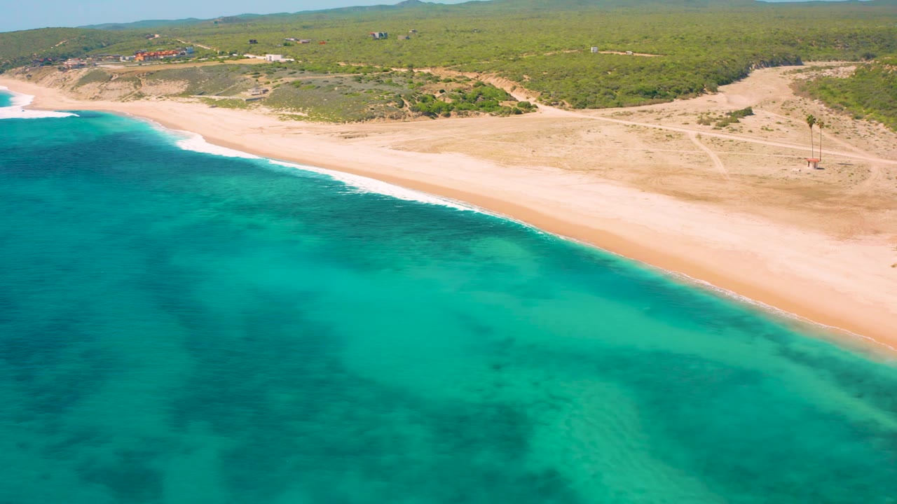 El Cardoncito aerial frame with bright turquoise sea in the foreground, pale sand across the middle, low green vegetation behind it, distant structures near the horizon, and no visible people.