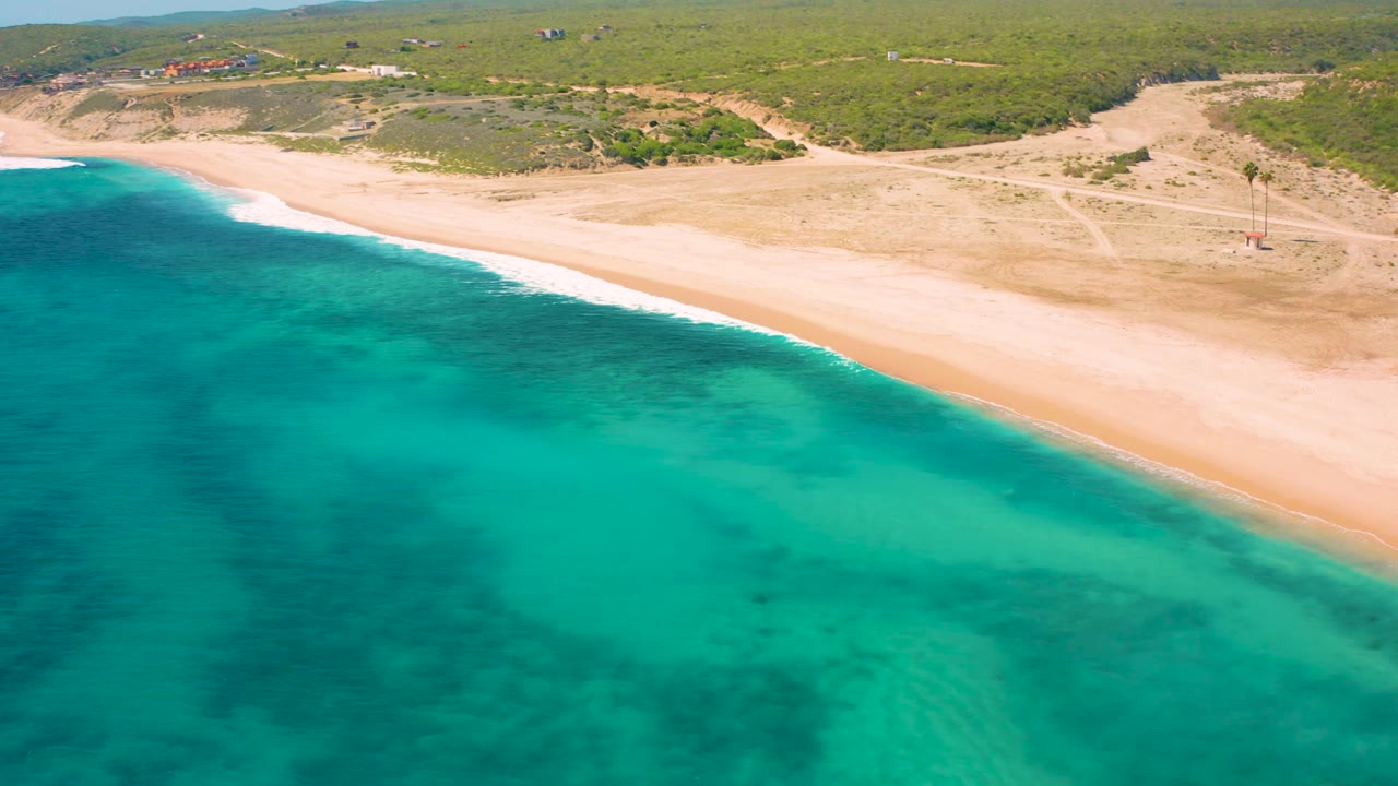 Aerial shoreline view with teal water, empty sand, inland tracks, and low vegetation.
