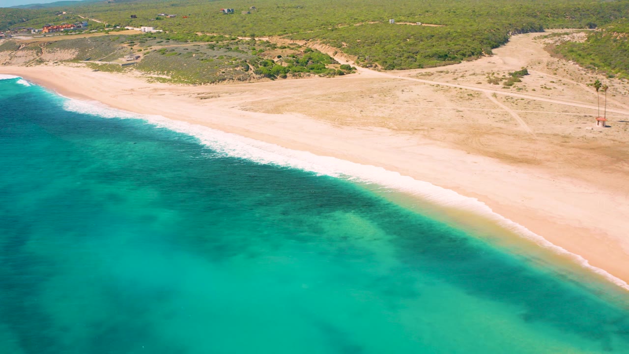 At 0:00, an elevated aerial view over El Cardoncito shows turquoise water in the foreground, pale sand along the shoreline, low green vegetation inland, and no visible people.