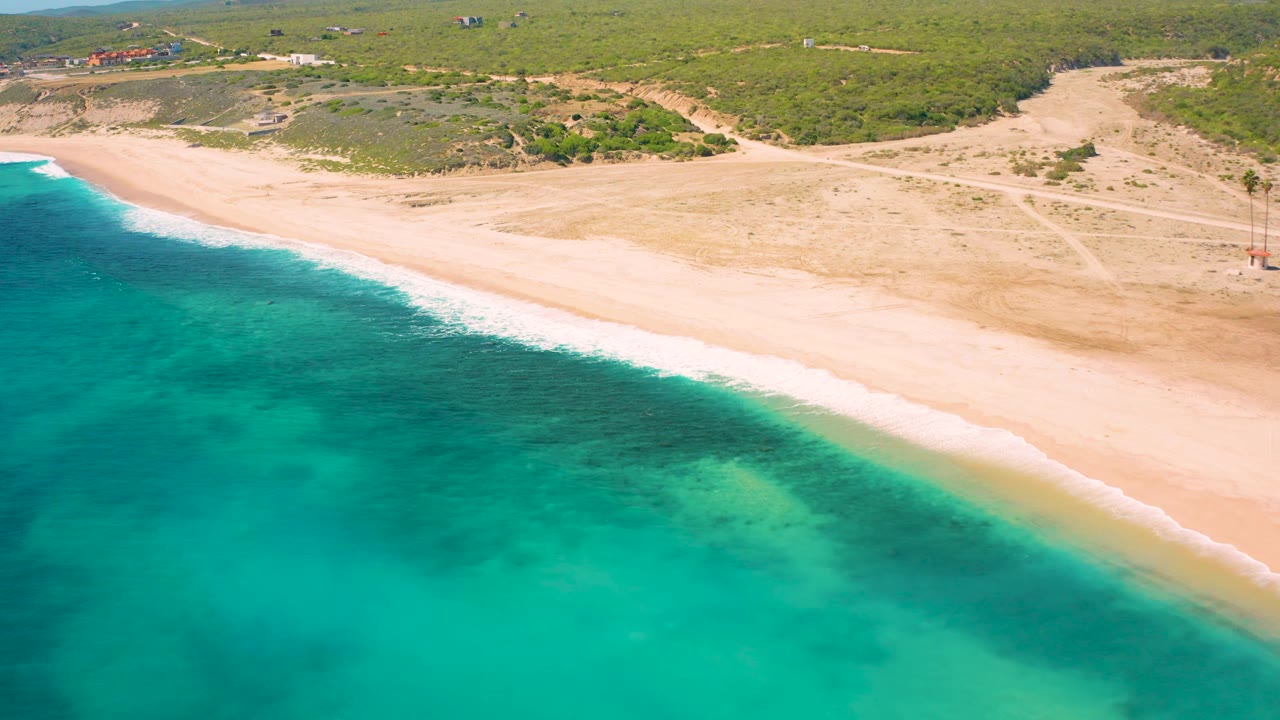 At 0:02, the El Cardoncito shoreline runs diagonally across the frame, with turquoise sea below, white wash at the beach edge, dry sand above it, inland scrub, and no visible people.