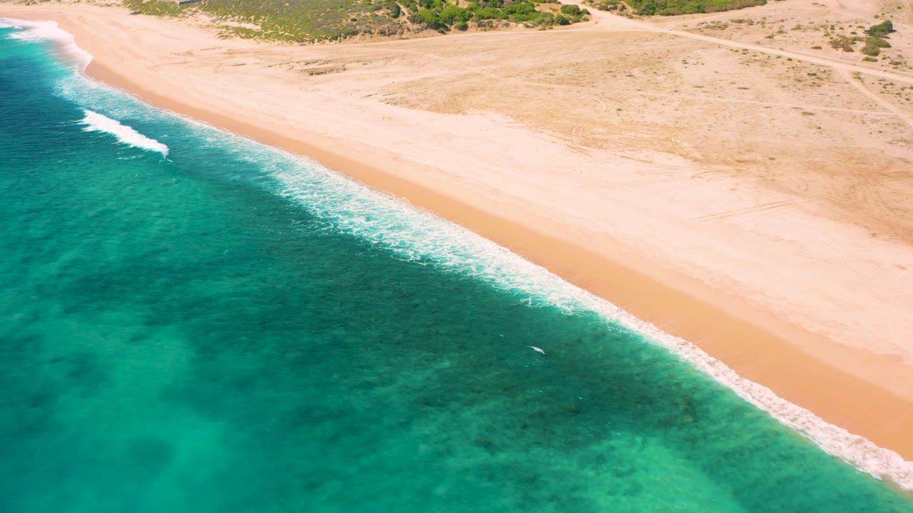 Aerial view over El Cardoncito, Baja California Sur, with turquoise water on the left, a diagonal band of surf, pale sand, scrub beyond the beach, and no visible people.