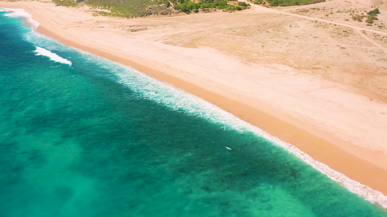 Overhead coastal frame in El Cardoncito showing a longer diagonal strip of white surf between teal water and pale sand, with dry scrubland at the top and no visible people.