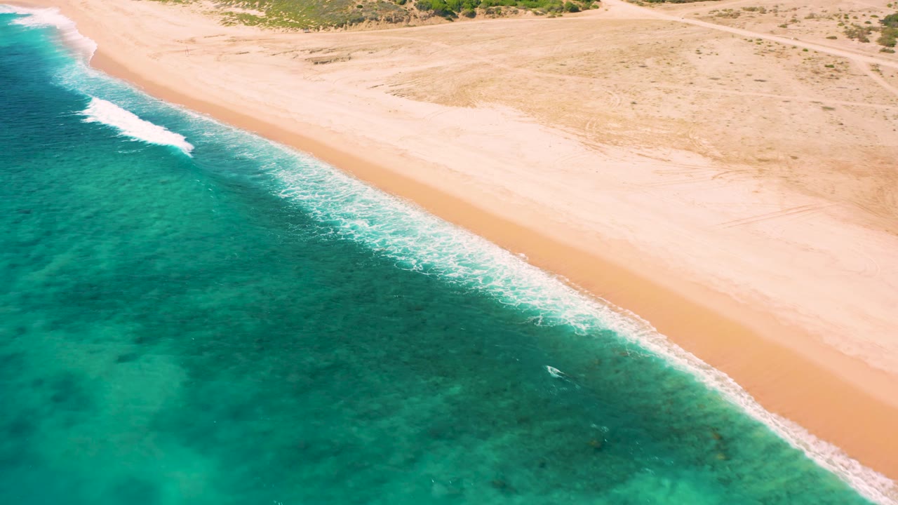 Aerial frame of El Cardoncito coastline with darker reef-like patches under turquoise water, white foam along the shore, open golden sand, and no visible people.