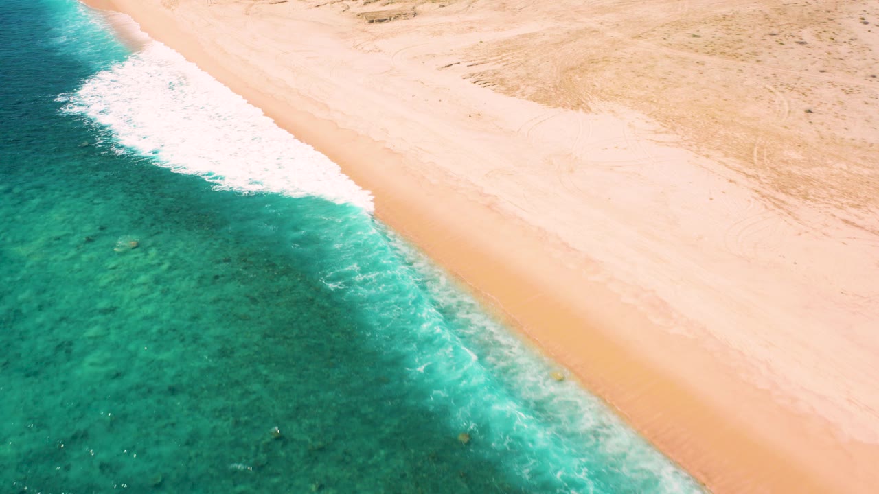 Aerial frame from El Cardoncito, Baja California Sur showing green water on the left, a diagonal strip of surf, pale sand on the right, and no visible people.