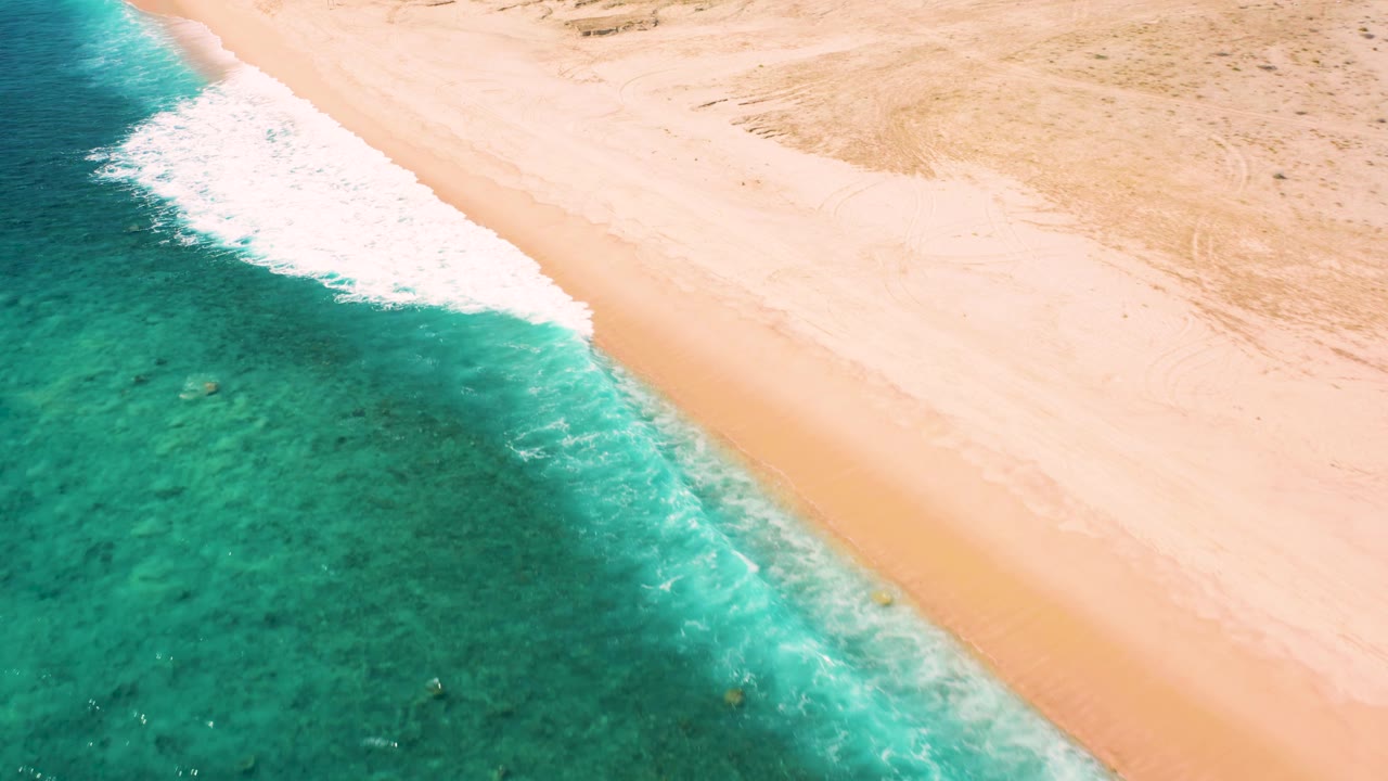 Aerial view in El Cardoncito, Baja California Sur with turquoise-green shallows, a thin white wave line, broad pale beach, and no visible people.