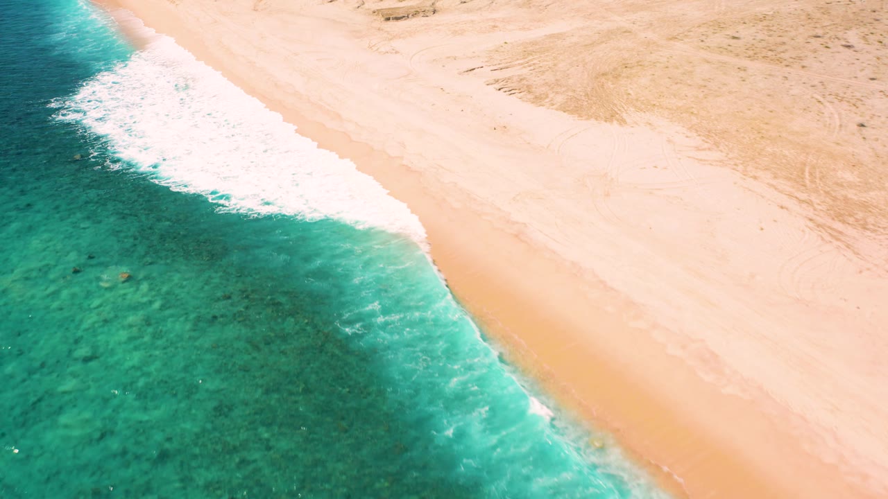 Aerial frame from El Cardoncito, Baja California Sur showing white foam spreading beside green water, dry sand beyond the tide line, and no visible people.
