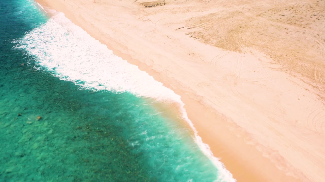 Drone-like overhead view in El Cardoncito, Baja California Sur with foamy surf curving across the center, darker green water at left, pale sand at right, and no visible people.