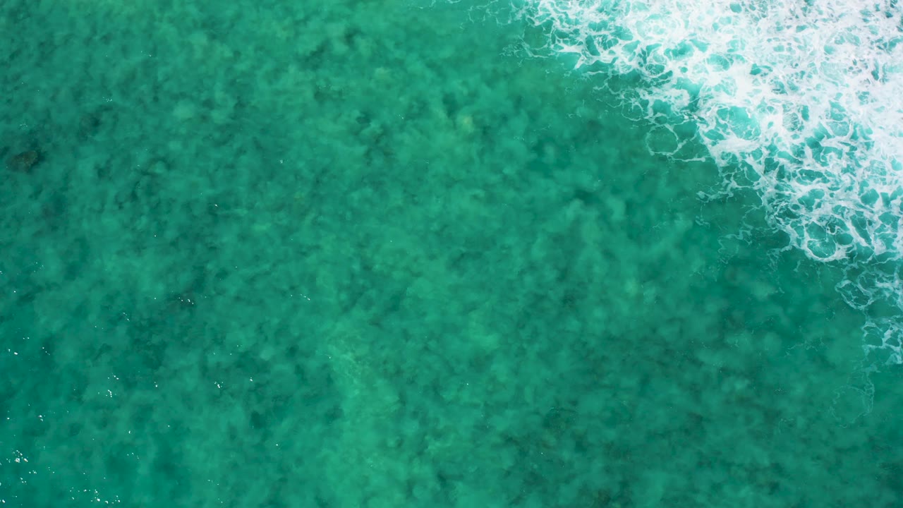 Top-down ocean frame from El Cardoncito with dense white foam at the upper right and open turquoise water with darker underwater texture below, no visible people.
