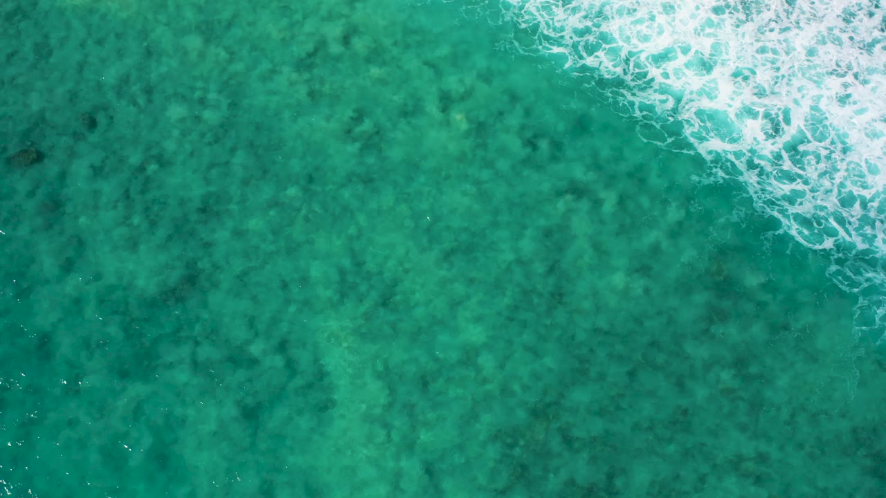 Overhead view of El Cardoncito water where white foam cuts diagonally into the turquoise surface from the upper right, with no land or visible people.