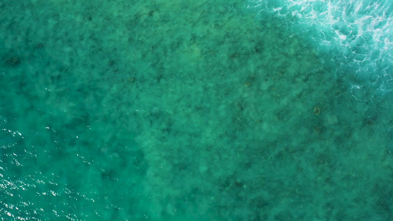 Aerial view of shallow turquoise water at El Cardoncito, Baja California Sur, with brighter white foam gathered along the upper edge and no visible people.