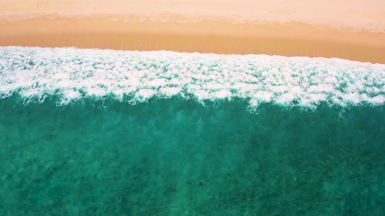 Aerial frame from El Cardoncito with a thick white foam line crossing the upper third and clear green-blue water below, with no visible people.