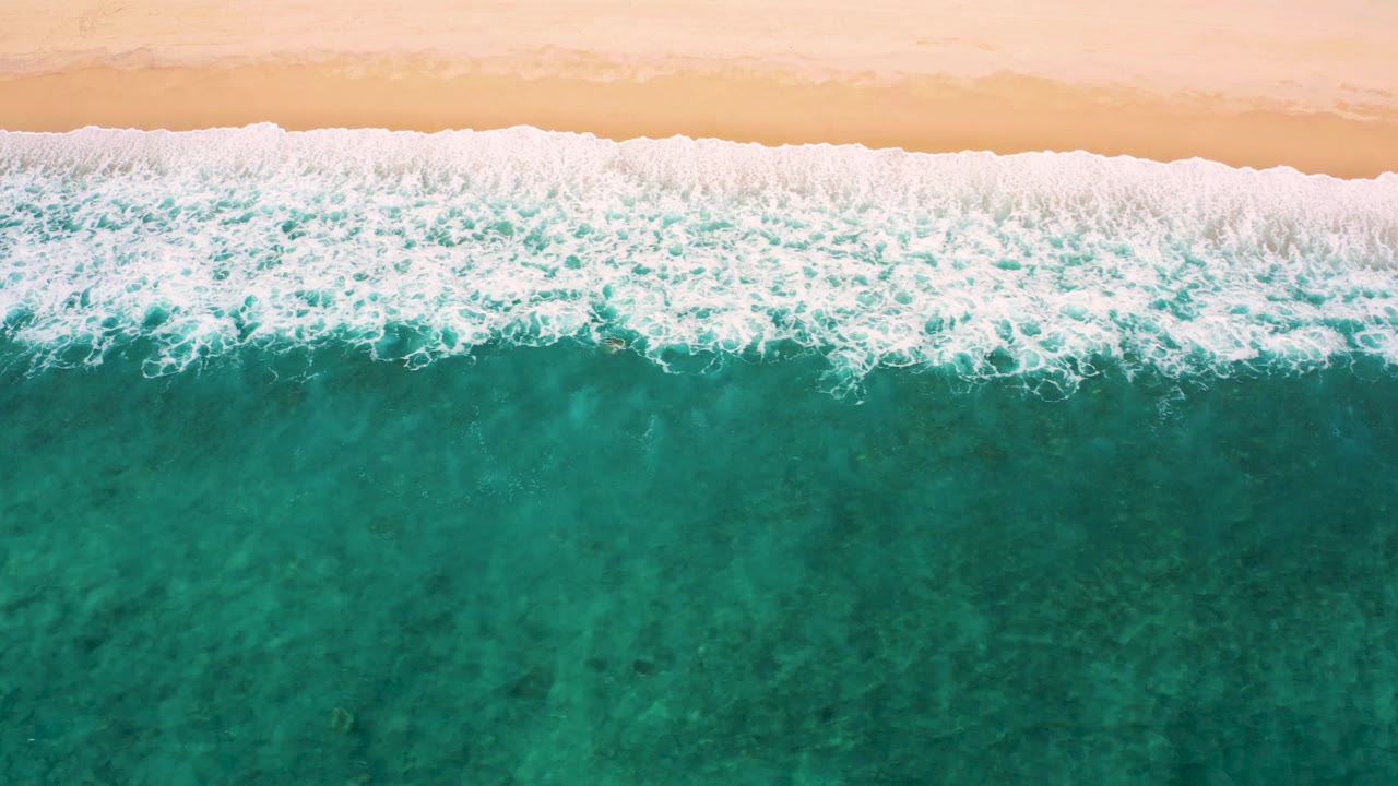 Overhead frame at El Cardoncito where a wider sheet of white surf reaches the pale beach while turquoise water fills the lower frame, with no visible people.