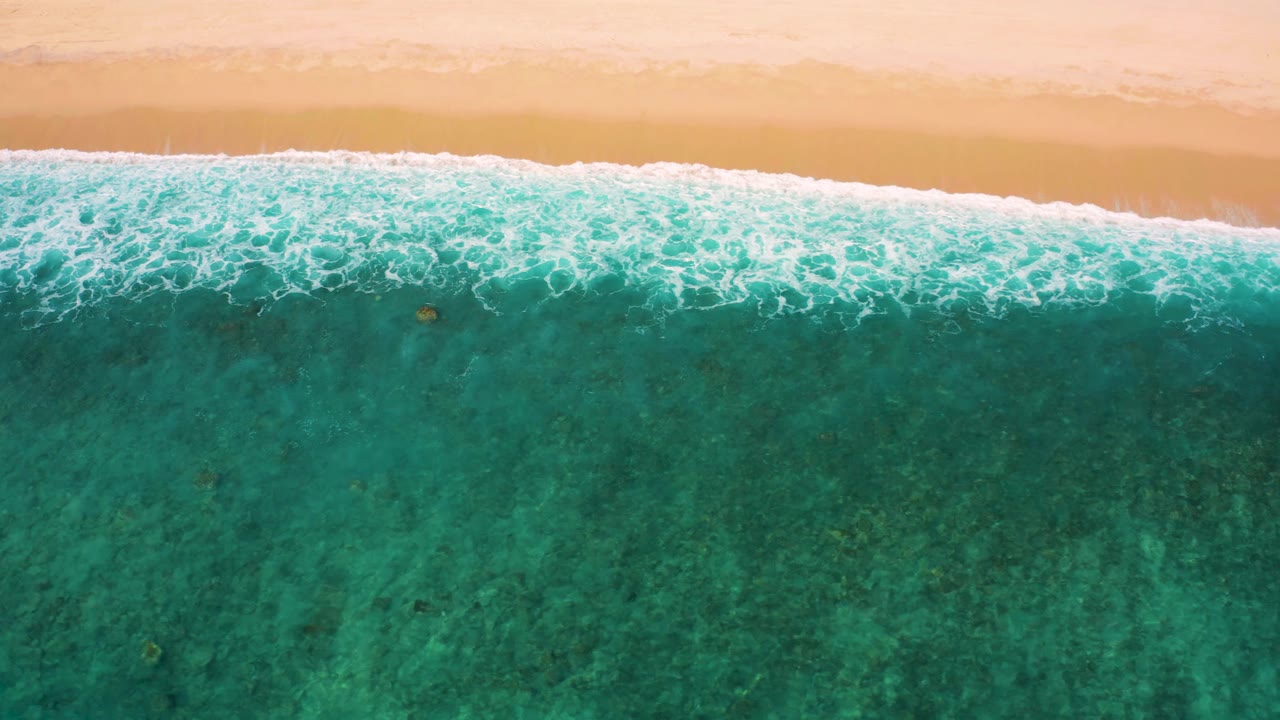 Aerial top-down frame at El Cardoncito shows a long white surf line beneath the sand, with blue-green water and mottled seabed texture, and no visible people.