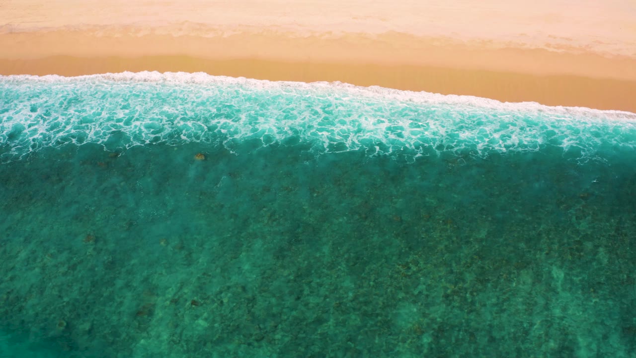 Top-down El Cardoncito beach frame with the foam line pushed close to the sand and a broad field of teal shallow water below, with no visible people.