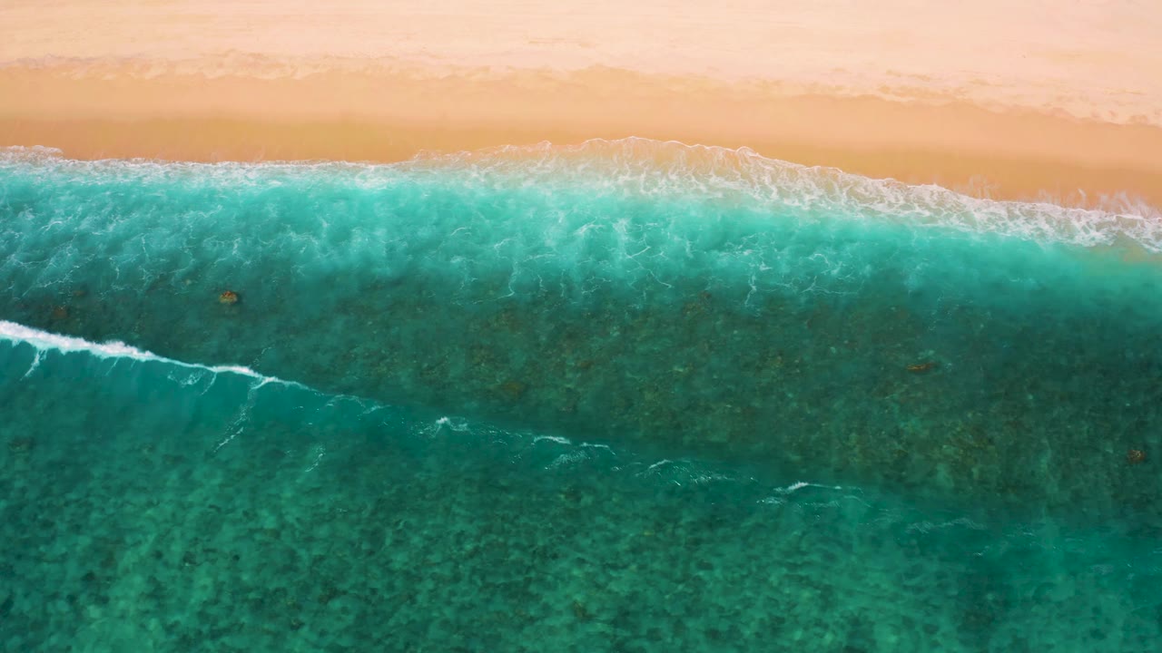 Aerial frame from El Cardoncito with clear green-blue water, pale beach along the top edge, and a narrow white wave beginning to break, with no visible people.
