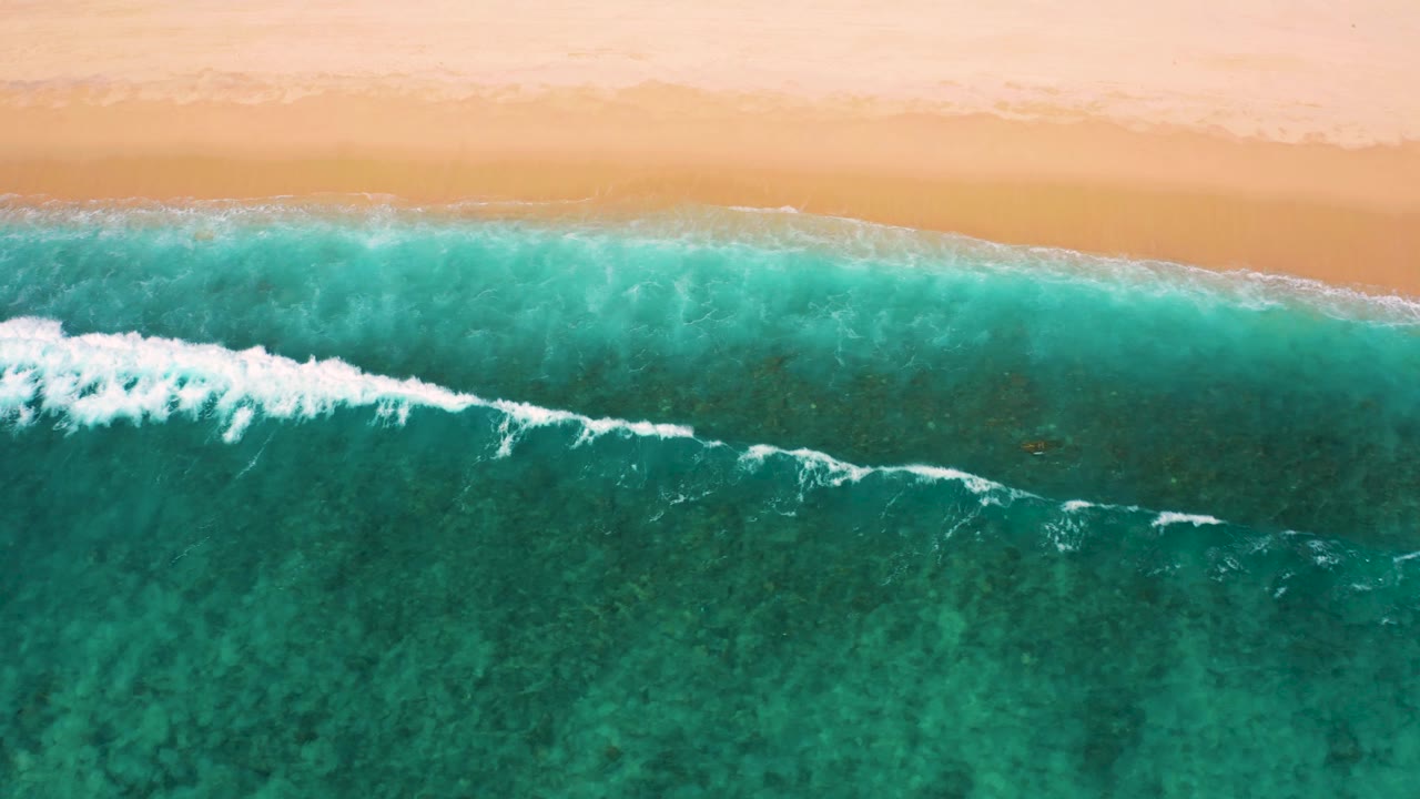 Aerial view at El Cardoncito where a breaking wave spreads white foam across green-blue water near the pale beach, with no visible people.