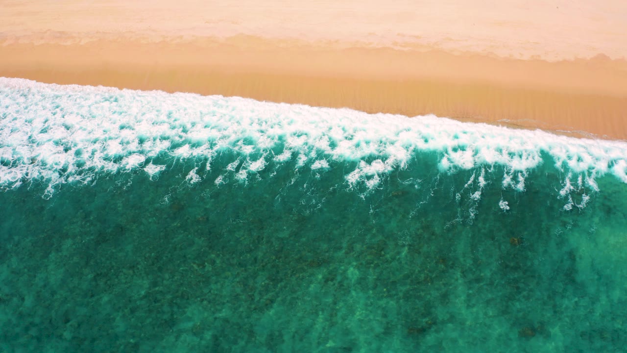Overhead aerial frame from El Cardoncito, Baja California Sur showing white surf foam along tan sand with teal green water below and no visible people.