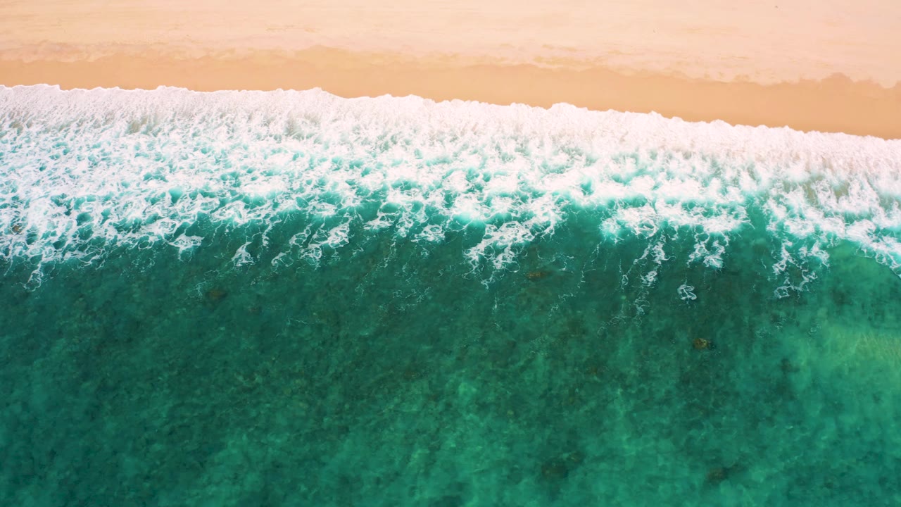 Aerial frame from El Cardoncito showing white water washing from the tan sand into teal green ocean, with foam curls and no visible people.