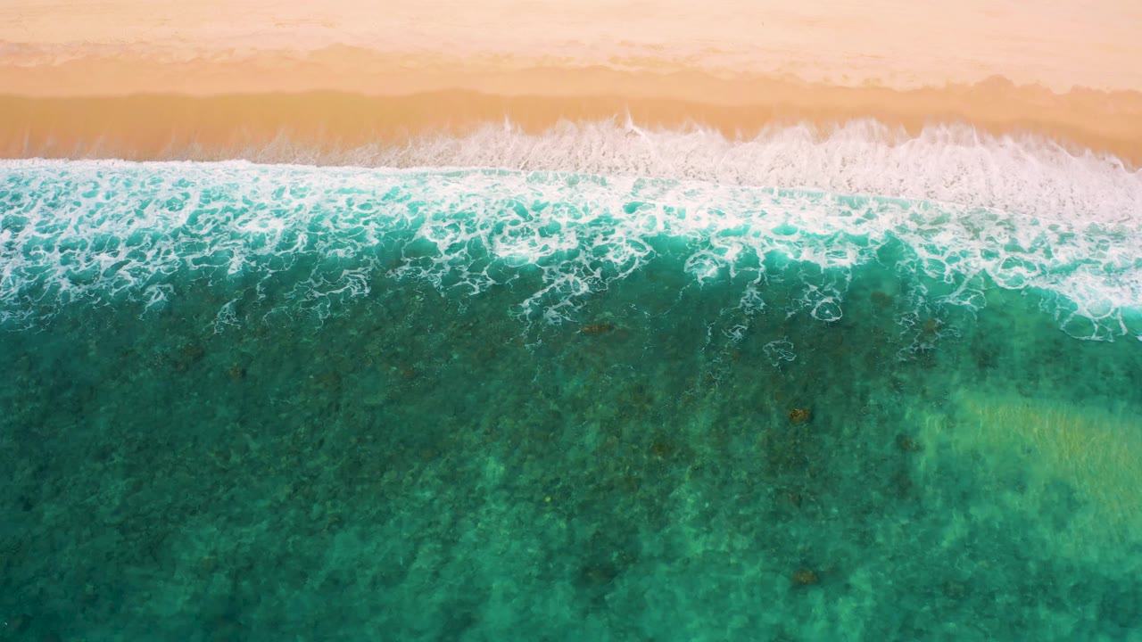 Aerial frame of white wave foam stretching across the sand-water boundary at El Cardoncito, with darker teal water below and no visible people.